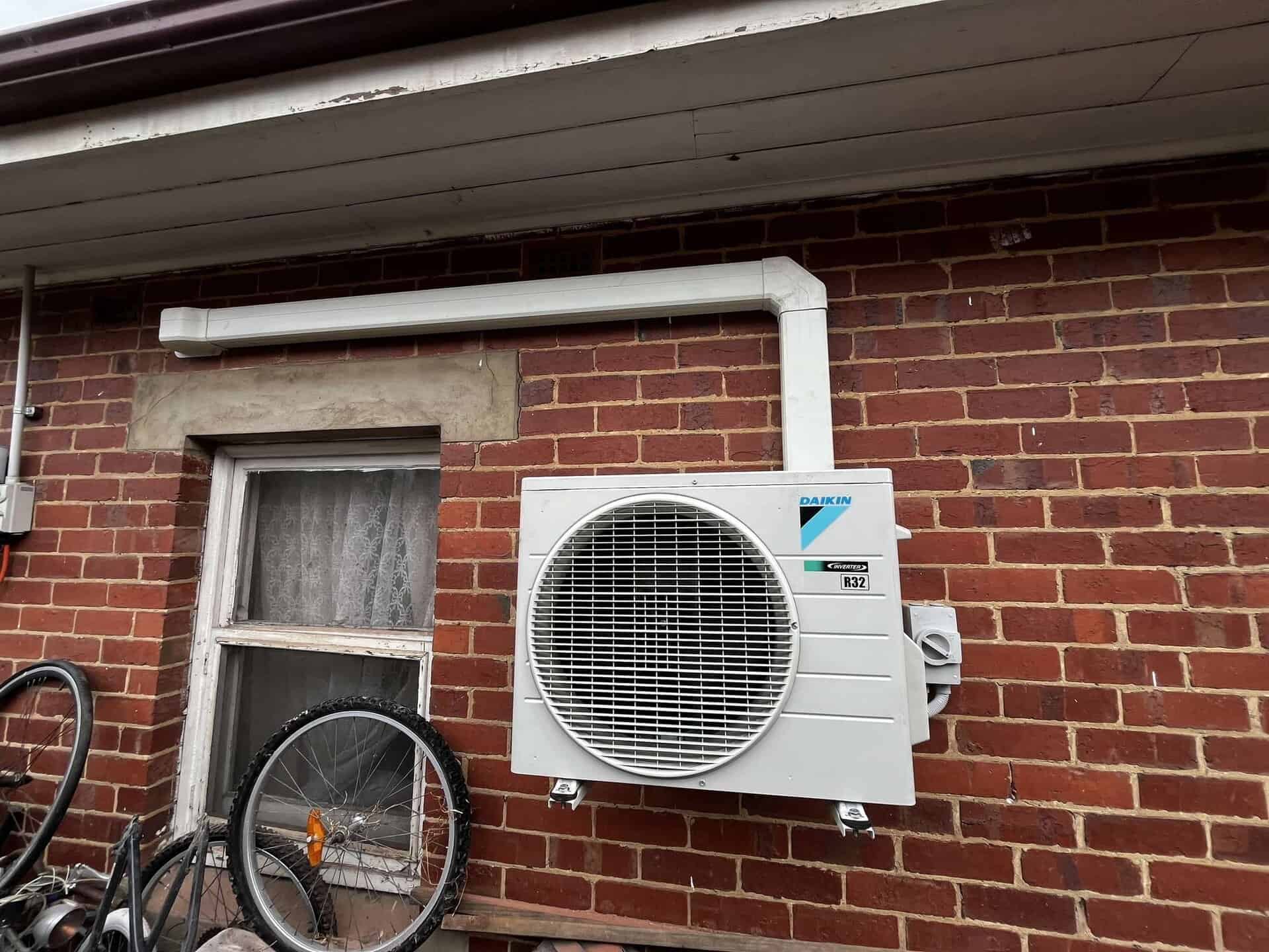 Bright white outdoor air conditioning unit mounted on a brick wall beside a small window, with a white pipe running horizontally above it outside a building.