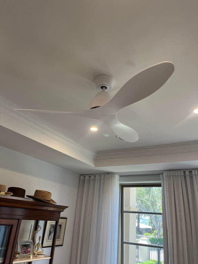 Modern white ceiling fan with three blades installed on a smooth, painted ceiling inside a living room, near a window with white curtains and decorative items on a wooden cabinet, natural light coming through the window, indoors in a residential setting.