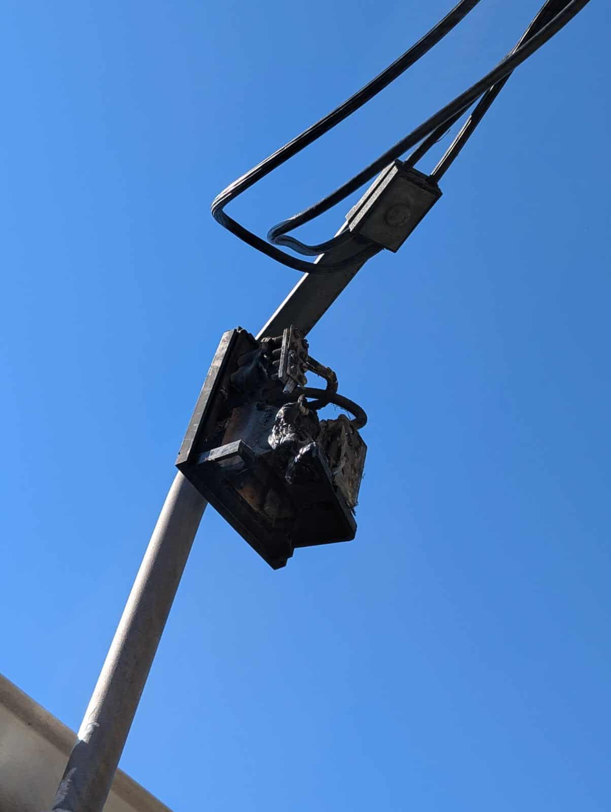 Damaged street light fixture on metal pole, mounted outside against a clear blue sky.